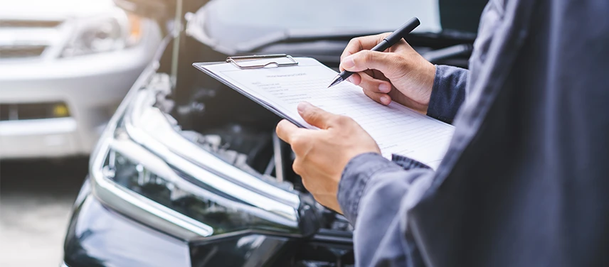 Professionnel de l’automobile remplissant une fiche de contrôle sur un porte-bloc devant un véhicule au capot ouvert, illustrant l’assurance RC Pro Auto.